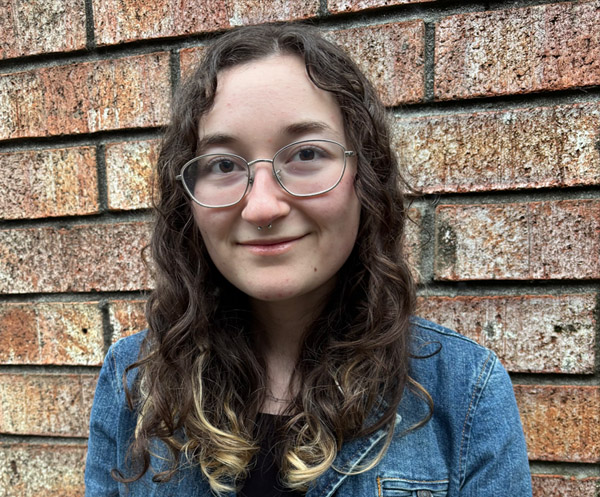 photo of Ashley - wearing blue jean jacket over black t-shirt. She's against a brick wall, looking at camera, long brown hair, glasses